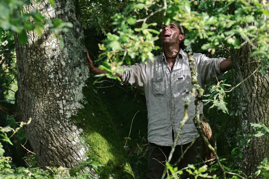 Un acteur dans les bois, bras ouverts, célébrant la nature lors du spectacle immersif 'De souvenirs en racines'. renforçant le lien émotionnel entre émerveillement, nature et art.