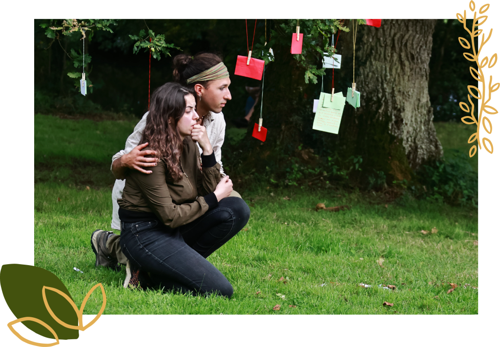 Un frere et une soeur à genoux dans l'herbe verte, entourées de souvenirs suspendues aux arbres, créant une atmosphère poétique et immersive.du spectacle de théâtre paysage De souvenirs en racine en in situ