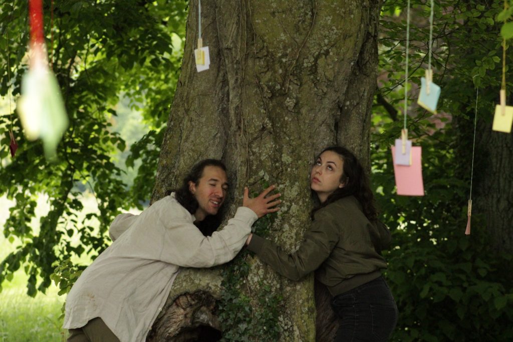 Deux actrices de théâtre enlacent un arbre couvert de papiers de souvenirs pendus aux branches, symbole de souvenirs et d'émotions et d'engagemebt écologique dans "De souvenirs en racines". Elles écoutent des arbres