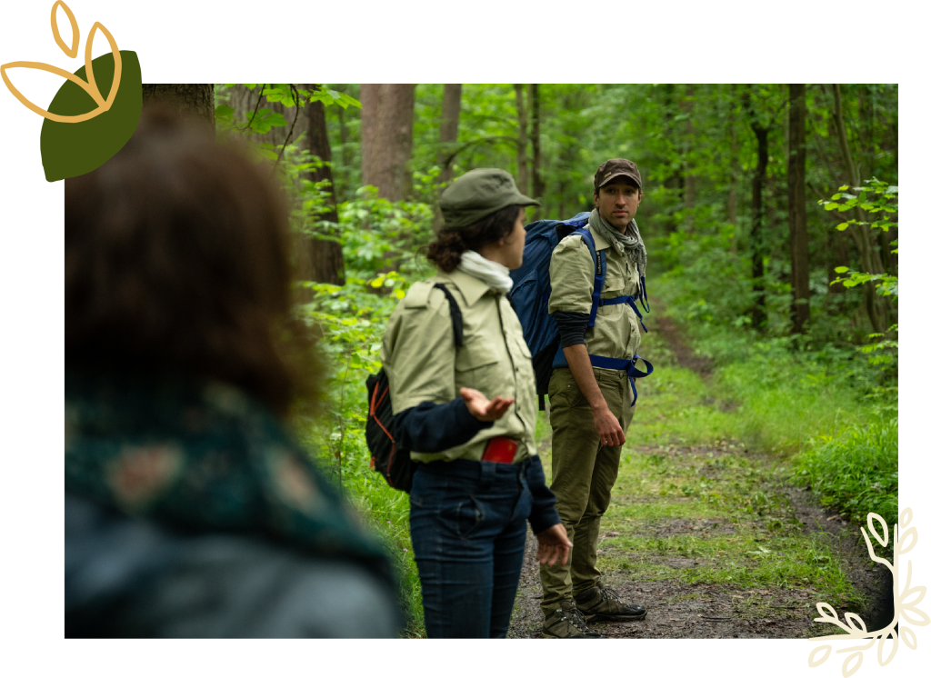 Deux gardes forestiers en pleine discussion dans une forêt verdoyante suivi du public. L'un d'eux se tourne vers ses compagnons, en désaccord sur la gestion forestière dans le cadre du spectacle déambulatoire de théâtre-paysage 'Les Écorcées si la forêt contait'