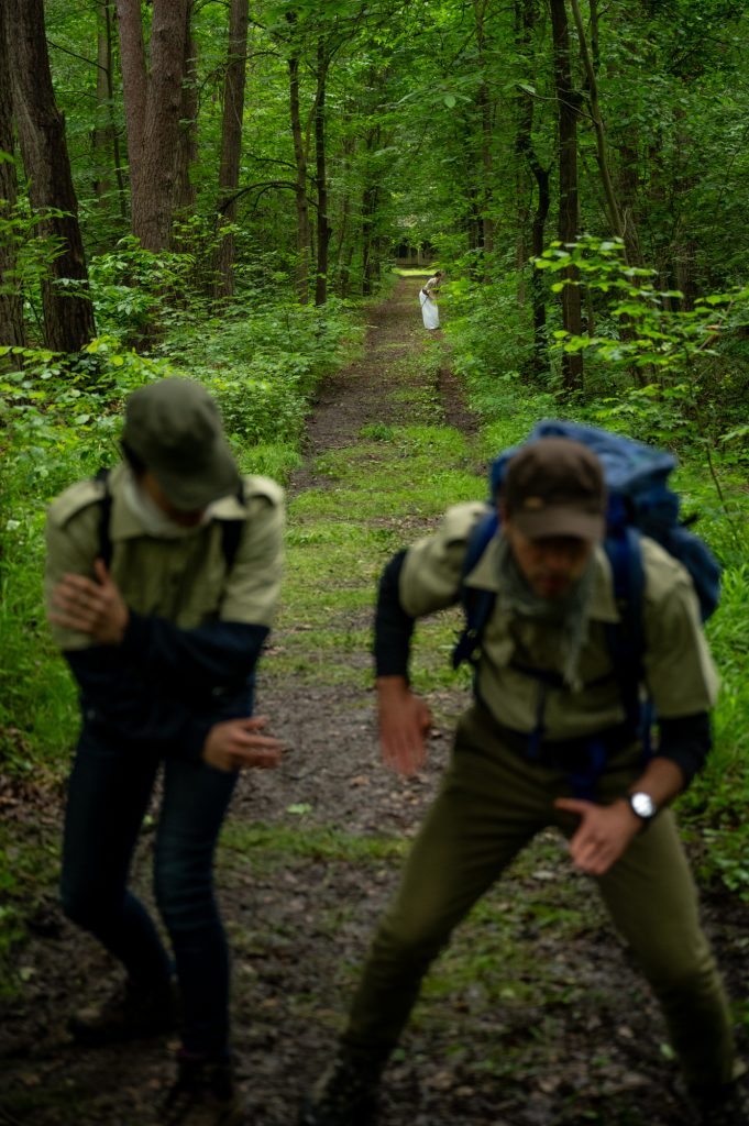 Deux gardes forestiers en pleine action lors du spectacle déambulatoire 'Les Écorcées', un projet immersif en forêt mêlant théâtre et nature. Une vieille femme en robe blanche se dessine au loin sur un chemin forestier, renforçant la dynamique de l'immersion