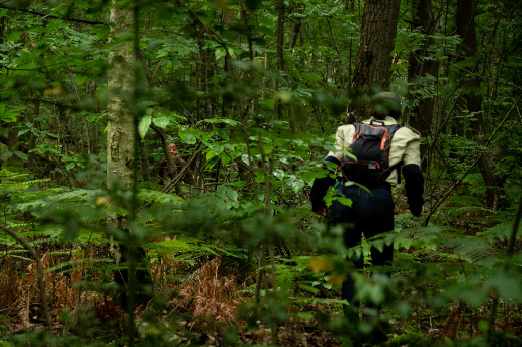 Un garde forestier en pleine forêt, marchant de dos en sous bois, un masque est visible au loin parmis les feuilles, symbolisant une connexion profonde avec la nature et la préservation des espaces boisés dans la déambulation 'les Ecorcées'.