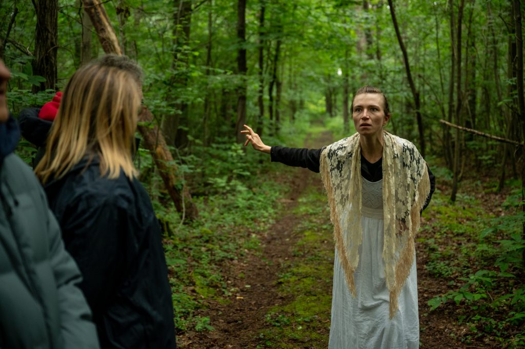 Une actrice vêtue d'une robe blanche interagit avec un membre du public lors d'une pièce de théâtre paysage en forêt. La rencontre se déroule dans le cadre immersif et éco-sensible de 'Les Écorcées, si la forêt contait', renforçant le lien entre nature et art