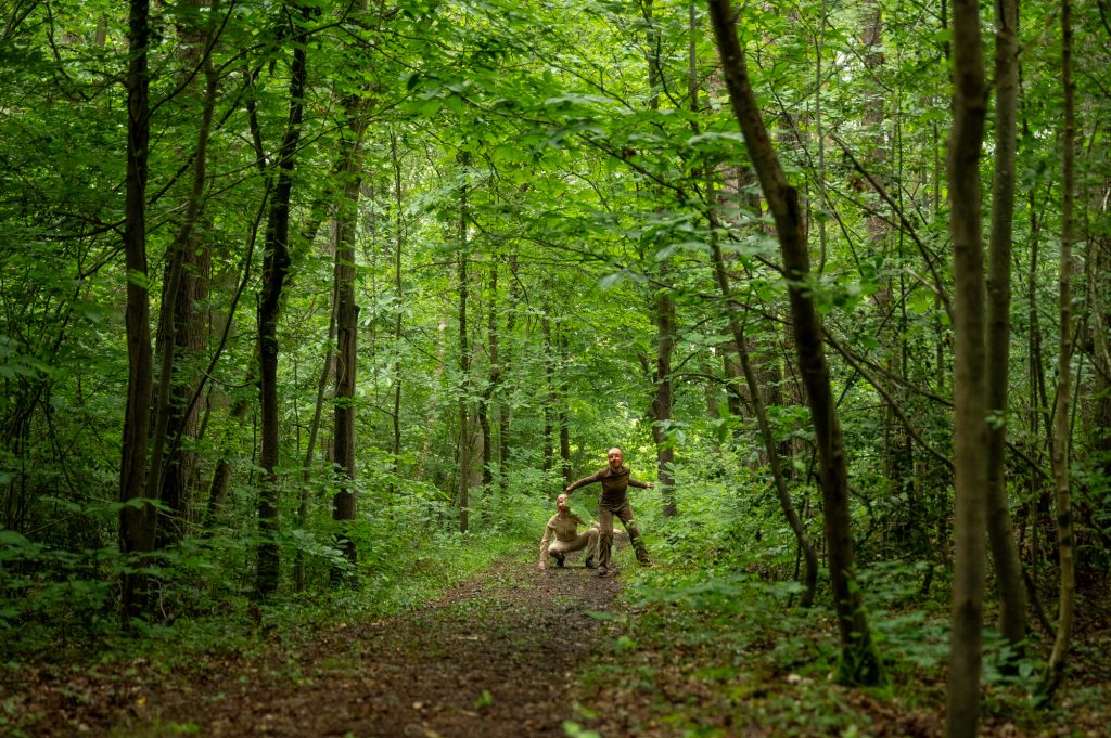 Deux acteurs masqués au cœur d'une forêt luxuriante, engagés dans une scène du spectacle immersif 'Les Écorcées'. Leurs postures dynamiques évoquent le mystère de leur présence et une tension palpable, contrastant avec le calme de la nature environnante.