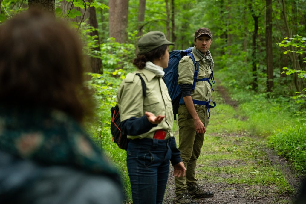 Deux gardes forestiers en pleine discussion dans une forêt verdoyante suivi du public. L'un d'eux se tourne vers ses compagnons, en désaccord sur la gestion forestière dans le cadre du spectacle déambulatoire de théâtre-paysage 'Les Écorcées si la forêt contait'